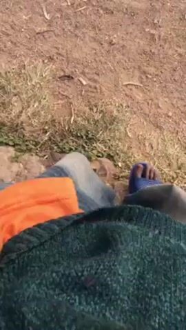 The video shows a first-person perspective of someone walking on dirt and grass. The person is wearing blue jeans, blue sandals, an orange shirt, and a dark green sweater. The ground is mostly dirt with some small patches of grass and twigs.