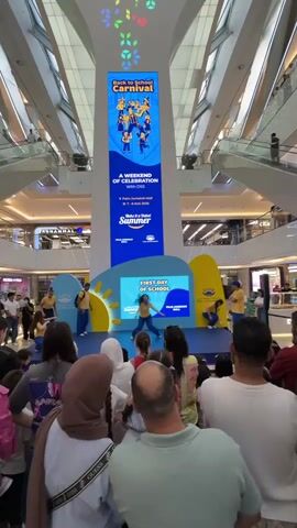 The video shows a "Back to School Carnival" event taking place inside a large, multi-level shopping mall. A large digital banner at the top advertises the carnival. Below this banner, a stage is set up with a smaller screen displaying "FIRST DAY OF SCHOOL" and graphics of a sun and clouds. Several young people in yellow shirts and blue shorts are performing on the stage, interacting with each other and the audience. A crowd of spectators, including adults and children, are gathered in front of the stage, watching the performance. The mall environment is visible in the background, with escalators and shops. The overall atmosphere appears festive and family-friendly.