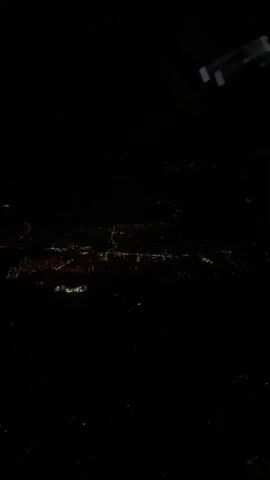 The video shows an aerial view of a city at night. The lights of the city are visible below, forming a grid-like pattern. The camera appears to be looking out of an airplane window, as a portion of the airplane's wing is visible in the upper right corner of the frame. The sky is dark, and there are no stars or moon visible. The city lights are predominantly orange and white, with some areas appearing brighter than others. The overall impression is of a vast urban landscape spread out beneath the darkness.