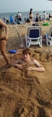 The video captures a sunny day at a crowded beach in Qinhuangdao, China. The main subject is a young person buried up to their chest in the sand, looking directly at the camera with a slightly surprised expression. Their arms are resting on the sand in front of them.

In the background, a bustling beach scene unfolds. Numerous people are visible, some walking along the shoreline, others sitting or lying on the sand. Beach chairs, a blue inflatable toy, and a green ring float suggest recreational activities. The ocean stretches out in the distance, with a gentle wave pattern visible. The sky is clear and the bright sunlight indicates it is likely daytime.

Other details include a pink pair of children's shoes lying in the sand near a wooden pole, and a blue toy. The overall atmosphere appears lively and typical of a popular beach destination. No visible text is present in the video.