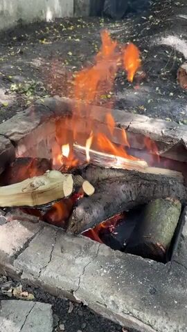 A close-up shot shows a brick fire pit filled with burning logs and flames. The fire pit appears to be outdoors, with dirt and scattered leaves on the ground surrounding it. The flames are bright orange and yellow, flickering and dancing around the wood. Some of the logs are partially charred, indicating they have been burning for some time. The fire pit itself is constructed from rough, gray bricks, with some of them appearing chipped or worn. The overall impression is one of a rustic, outdoor setting where a fire is being maintained.