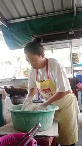 A woman with her hair pulled back in a bun, wearing a white t-shirt and a yellow apron, is standing and mixing white powder in a large green basin with her hands. The woman appears focused on her task. The scene seems to be an outdoor food stall or preparation area, with a metal roof overhead, some cooking equipment visible in the background, and various containers and utensils scattered around.