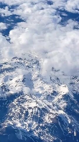The video captures an aerial view of a vast, snow-covered mountain range, likely the Dinaric Alps given the context of Seljani, Montenegro. White, fluffy clouds partially obscure the jagged peaks, with some clouds floating above and others clinging to the mountainsides. Patches of blue sky are visible between the clouds, suggesting a clear day at high altitude. The bright sunlight reflects off the pristine snow, creating strong contrasts and shadows that highlight the rugged terrain. The view appears to be from an airplane, as indicated by the high vantage point and the subtle movement. The sound of an airplane engine hums softly throughout the recording. There are no people, visible objects other than the natural landscape and clouds, or discernible text.