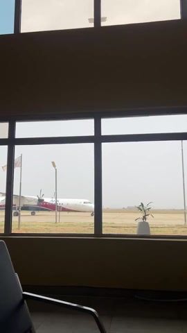 The video shows a white and red propeller plane on a runway, viewed through the window of a waiting area. The plane is stationary on the tarmac, with a flag and light pole visible nearby. Inside the waiting area, a gray chair sits near the window, and a small potted plant is visible outside. The day appears hazy or overcast.