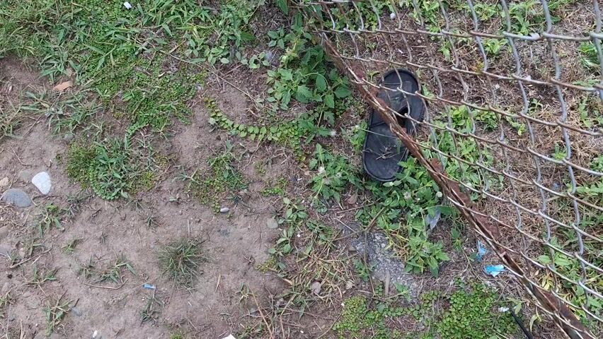 The video opens with a ground-level view of an outdoor scene, featuring a patch of dirt and sparse green grass. To the right, a rusty chain-link fence runs horizontally across the frame. Lying on the ground near the fence is a single black sandal. As the video progresses, a person's feet, clad in grey and black sneakers and black pants, enter the frame from the left and stand on the dirt path. The camera slowly pans slightly to the left and upward, revealing more of the person's legs and shoes. The ground around them is a mix of dry earth and scattered weeds and grass. Small white pebbles are visible on the dirt. The overall setting appears to be an unpaved area, possibly a path or roadside, in Panigah, Indonesia, during what seems to be daylight hours. No specific activities other than standing are depicted, and there is no visible text.