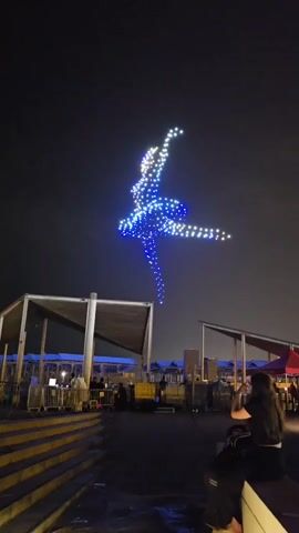 The video shows a drone light show at night. A swarm of blue and white lights in the sky forms the shape of a dancing ballerina. The drone formation is suspended in the dark sky above a low-light outdoor venue with structures and people visible in the foreground. A person is seated on the right, facing away from the camera, and appears to be taking a photo or video of the display.
