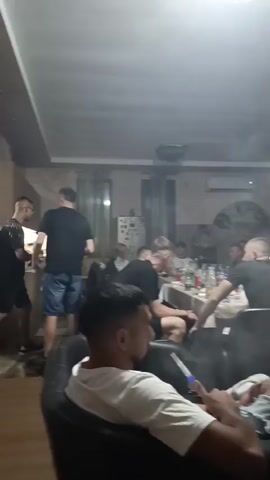 A group of men are gathered indoors, some seated at a table with food and drinks, while others are lounging on sofas. Several people are smoking from shishas, and there is a noticeable amount of smoke in the air. The room appears to be a living space with an air conditioner visible. One man in the foreground is relaxing on a couch, holding a shisha pipe. The overall atmosphere suggests a casual social gathering.