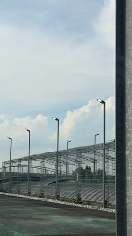 A vertical, outdoor, medium shot captures a cloudy, blue sky with large, white, puffy clouds. In the lower half of the frame, a large, industrial structure with a metal framework and railings stretches horizontally. Several tall, modern streetlights with LED fixtures are positioned along the top of this structure. The ground in the foreground is dark and appears to be paved. The image is static and appears to be a still frame from a video.
