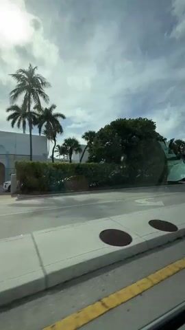 The video shows a view from inside a moving vehicle looking out at a street scene. On the right side of the frame, there is a large green and yellow structure, possibly a boat or a large decorative piece, parked on the side of the road. Further down the road, there is a modern-looking building with large windows and columns, surrounded by palm trees and lush greenery. Several vehicles are visible, including a white pickup truck and a dark-colored SUV. The road has clear lane markings, and there are hedges and flower beds along the side of the street. The sky is partly cloudy with bright sunlight. The camera appears to be handheld or mounted in the vehicle, resulting in some slight movement and occasional reflections on the window.