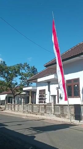 A red and white flag on a flagpole flies against a clear blue sky. The flag is positioned in front of a white house with a brown roof. The house has windows and a fence in front of it. There is a tree to the left of the house. The flag appears to be Indonesian. The ground in front of the house is paved, and there are shadows cast on the ground.
