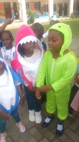 The video shows a group of young children in costumes, likely at a school or event. Several children are dressed as sea creatures, with one in a blue shark costume and another in a pink shark costume. Another child is wearing a green dinosaur costume. A woman, possibly a teacher, is visible in the background. The children appear to be standing in a line or gathered together, with some engaging with the camera. The environment appears to be outdoors, possibly a courtyard or playground, with a building and trees in the background. The lighting suggests it is daytime. The video is shaky and the focus shifts between different children. In the latter part of the video, the costumes change to superhero and princess attire, indicating a potential costume change or a different group of children.