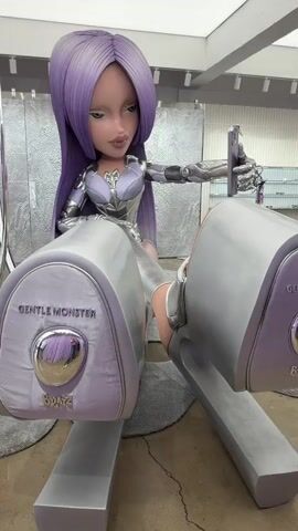 The video shows a life-size, seated, fashion doll with purple hair and silver clothing, seemingly a promotional collaboration between Gentle Monster and Bratz. The doll, with its oversized head and exaggerated features typical of Bratz dolls, sits with her legs spread, each foot resting on a large, cylindrical footrest bearing the "Gentle Monster" and "Bratz" logos. The footrests are silver with pale lavender accents. In her right hand, the doll holds a silver phone, presumably taking a selfie. The doll's clothing appears to be a stylized, futuristic design. The environment is a modern retail space with silver display elements, shelves holding eyewear, and a carpet. People can be seen in the background moving around. The lighting in the space is bright and even.