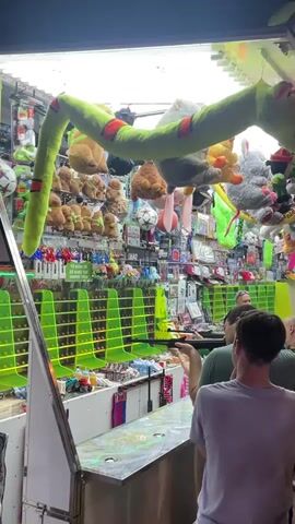 The video shows a carnival game booth at night. The booth is filled with prizes, including stuffed animals and toys. Two young men are playing the game. The game involves shooting at targets with a toy gun. The first man is standing at the booth, aiming the gun at the targets. The second man is standing behind him, watching. The game booth is brightly lit, and there are people in the background. The atmosphere is lively and festive.