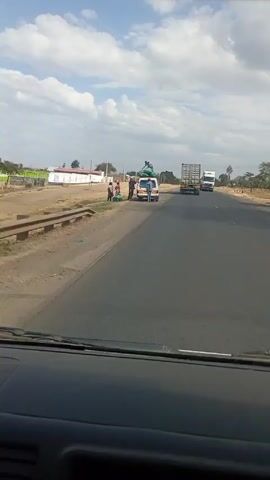 The video shows a view from inside a vehicle driving on a highway on a sunny day. In the foreground, the dashboard is visible. On the side of the road, a white and yellow van with people loading items onto its roof is stationary. The road has two lanes going in each direction. Two trucks are visible ahead, one carrying a container and the other a tanker. In the background, there are scattered buildings and trees under a sky with patchy clouds. As the vehicle moves forward, it passes the van and the trucks are seen moving in the opposite direction. There are a few people walking along the side of the road in the distance.