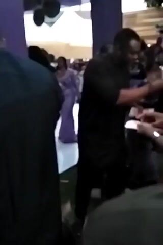 The video shows a scene indoors, possibly at an event. Several people are present, including one in a maroon robe and cap, holding a staff. There are also individuals dressed in black and a woman in a long purple dress. People appear to be handing out white items, possibly money or tickets. The background includes purple pillars and a white floor. The overall impression is a gathering with possible ceremonial or celebratory aspects. The video is a bit shaky and the view is partially obstructed by people in the foreground.