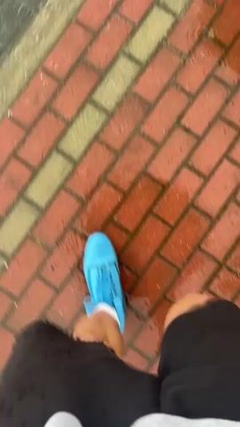 A person wearing blue shoe covers and black shorts walks on a wet brick and asphalt sidewalk, with water puddles visible on the ground. The person is walking towards a crosswalk with white stripes. The camera angle is from a high perspective looking down at the person's legs and feet. The overall scene suggests it is raining or has recently rained.