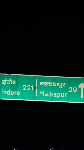 A green road sign displays text in both Hindi and English. The Hindi text reads "इंदौर" and "मलकापुर", with the number "221" between them. The English text reads "Indore" and "Malkapur", also with "221" between them. The sign appears to be lit from behind, and the background is dark, suggesting nighttime. The video then transitions to a shot of a dark road at night, illuminated by streetlights. Several streetlights are visible in a line, and there are also some red and yellow lights, possibly from vehicles or other signs. The overall scene is dark and atmospheric, with a sense of driving through a city at night.