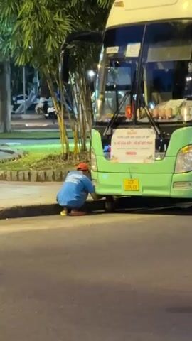 A person wearing a blue t-shirt and red hat crouches next to the rear wheel of a green bus. The person appears to be working on the underside of the bus. The bus has a license plate that reads "60F 005.68". There is a sign on the back of the bus with text in Vietnamese. The scene appears to be outdoors, possibly at night, with some trees and foliage in the background.