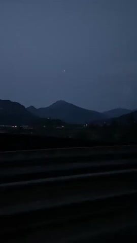 This is a vertical video taken from a moving vehicle at dusk. The foreground consists of dark horizontal lines, likely guardrails or barriers of a road. In the mid-ground, silhouetted trees and buildings are visible, along with faint lights from a settlement. The background is dominated by the dark shapes of mountains under a dusky blue sky. A single bright star or planet is visible in the upper portion of the sky. The video captures a sense of transit through a landscape as daylight fades.