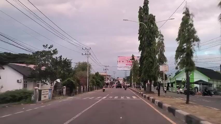 The video shows a road view from a moving vehicle on what appears to be a street in an urban or semi-urban area. The street has power lines overhead and trees lining the roadside. There is a large billboard visible with an advertisement that includes images of people, most likely for a local campaign or event. Several motorcycles and a car are visible moving on the road. Buildings and houses line the street, along with a fenced area on the left side of the road. The weather looks overcast, with a slightly pinkish hue to the sky. The road is marked with a crosswalk, and black and white markers line the side of the road. The overall scene suggests a typical day in a small town or suburban area.
