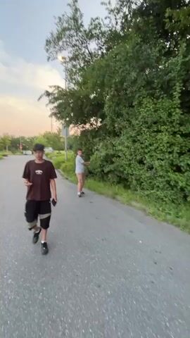 This media file shows a street scene in Oelde, Germany, likely in the late afternoon or early evening given the soft, warm light and pastel colors of the sky. Two young males are the primary subjects, engaged in casual activity.

In the foreground of the first clip, a young male wearing a brown t-shirt, black shorts with knee pads, and black sneakers walks towards the camera while holding a phone. His expression is neutral, and he appears focused on his device. Behind him, another young male in a light blue t-shirt and plaid shorts walks away from the camera.

The subsequent clips focus on a third young male in a bright pink t-shirt and matching pink shorts, also wearing sneakers. He is holding a phone in his right hand, seemingly filming himself or taking a selfie, and holding a dark object, possibly a cigarette or vape pen, to his mouth with his left hand. He is walking along a paved path, with lush green trees and bushes lining the right side of the path. In the background, streetlights and trees indicate an urban or suburban setting. The overall mood is relaxed and informal, suggesting a casual outing or a moment captured during outdoor recreation.