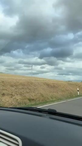 The video shows a view from inside a vehicle driving on a paved road. The road is bordered by a grassy embankment and trees. In the distance, a wind turbine is visible against a cloudy sky. A white and black striped post is seen on the side of the road. The vehicle appears to be moving forward.