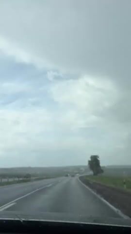 The video shows a car driving on a wet, two-lane highway on a cloudy day. Rain is falling, and the car's windshield wipers are in use. Several other vehicles are visible on the road ahead. Green fields line the side of the road, with trees and some buildings visible in the distance. The sky is overcast with a mix of white and dark grey clouds. The video is shot from the driver's perspective.