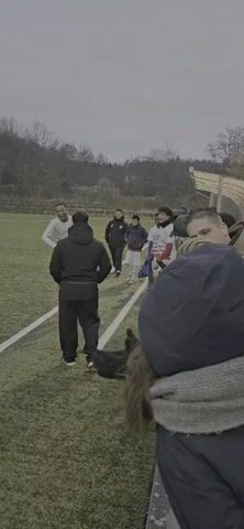 The video shows a soccer player running along the white sideline of a soccer field during a game. The player is wearing a white uniform with the number 16 on the back. He is running towards the goal in the distance. There are other players on the field, some wearing yellow uniforms. Several people are standing on the side of the field, watching the game. The environment is a green field with trees and a light post visible in the background. The lighting suggests it is either dusk or an overcast day. The camera is positioned near the sideline, following the player's movement.