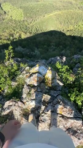 The video captures a high-angle, first-person view from the edge of a rocky precipice overlooking a vast, lush landscape. The viewer's own feet, clad in neutral-toned hiking boots, and the bottom of their light-colored pants are visible on the rocky terrain.  The rocks are scattered with small green bushes. The valley is filled with dense, green forest. The shadow of the person filming stretches down the mountainside, indicating a strong light source.  In the distance, flat plains are punctuated with clusters of buildings. The sky is clear blue. The camera angle gradually elevates, revealing more of the landscape. A strong wind is present, based on the audio.