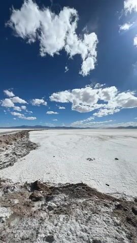 The video captures a wide-angle view of a vast, white salt flat under a bright blue sky dotted with white, fluffy clouds. The sun is shining intensely, casting lens flares and creating a bright, almost washed-out appearance in the sky. The foreground features a rough, dark, and textured embankment, possibly made of dirt and salt deposits, which gives way to the expansive, cracked white surface of the salt flat. In the distance, a faint, hazy line of mountains or hills can be seen on the horizon, suggesting the immense scale of the landscape. The overall environment appears arid and desolate, characteristic of a desert or salt pan. There are no people or animals visible in the video. The camera pans slowly to the right, revealing more of the salt flat and its textured surface.
