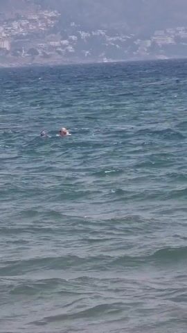 The video shows a wide shot of a body of water with waves. In the distance, there is a shoreline with buildings and trees on a hill. In the middle of the water, there are two people. One person is floating on an inflatable raft, appearing to be a woman with blonde hair wearing a bikini. Another person, possibly a man, is swimming in the water to the left of the raft. The water is a deep blue-green color and appears somewhat choppy. The sky is overcast.