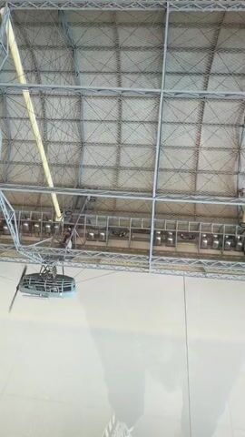 A drone with a blue pod and a spinning propeller flies horizontally across an ice rink. The drone is suspended from a wire that is attached to a grid-like ceiling structure. Above the ice, there is a row of caged areas with people lying down. The ceiling is covered with a netting material and supported by yellow beams. The drone is casting a shadow on the ice.