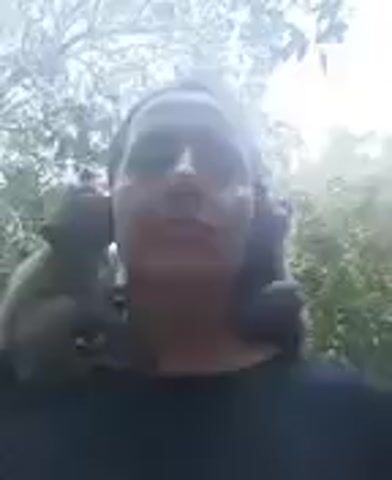 A person is filmed outdoors in what appears to be a natural, heavily vegetated environment, possibly a forest or jungle in Ban Tai, Thailand. The bright sunlight filters through the leaves, creating a glowing effect in the background and overexposing parts of the scene. The subject is a person, likely with dark hair, which might be styled in pigtails or two buns, and they are wearing dark clothing. Their face is largely obscured by the bright light and shadows, making it difficult to discern their features or expression. The person appears to be stationary, looking towards the camera. No specific activities or objects are clearly visible beyond the person and the natural surroundings. The time of day is clearly daytime, given the strong sunlight.