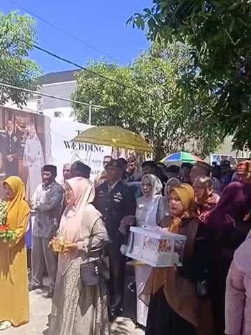 A group of people are gathered outdoors for a wedding celebration. A large banner in the background features a photo of a bride and groom and the words "THE WEDDING BE HAPPY". In the foreground, several women are dressed in traditional clothing and headscarves. One woman on the left holds a bouquet of flowers. Another woman in the center holds a small golden object, possibly a gift. A woman in the foreground right holds a clear plastic container with a white object inside, likely a cake or gift. A man in a military uniform stands among the guests. The environment appears to be a street lined with trees and buildings, under a clear blue sky. Some guests are holding umbrellas to shield themselves from the sun. The overall atmosphere suggests a joyous occasion.