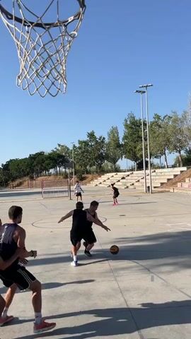 A group of young men are playing basketball on an outdoor court. The camera is positioned beneath the basketball hoop, looking up at the players and the sky. The court is concrete with painted lines. In the background, there are trees, stadium seating, and streetlights. The men are wearing athletic clothing, and some are shirtless. The video captures a moment during the game, with one player attempting to shoot the ball while others observe. The ball goes through the hoop.