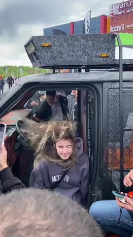 A young woman with long blonde hair is sitting in the open door of a modified black vehicle, possibly a jeep or an SUV, with her hair being blown by the wind. She is wearing a dark hoodie with white text. A man in a black jacket and white t-shirt is standing behind her inside the vehicle. Several other people are visible around the vehicle, with some holding up phones, seemingly recording or taking pictures. The vehicle has a rugged, customized exterior with metal plating and lights on top. In the background, there is a large building with retail signs, including "Kari" and "Sportmaster". The overall scene appears to be an outdoor event or gathering.