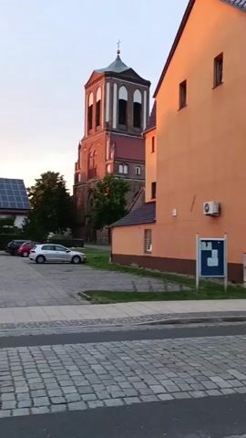 This video captures a street view in Gartz (Oder), Germany, likely during the late afternoon or early evening as indicated by the warm, soft lighting. The scene is dominated by charming, European-style architecture. On the left, a distinctive brick church tower with a green-capped steeple stands prominently against a pale, cloudy sky. Next to it, a modern building with solar panels suggests a blend of old and new in the town.

The foreground features a cobblestone street with a modern asphalt strip running down the center, characteristic of many European towns. Several cars are parked in a lot adjacent to the buildings, including a silver sedan and a red hatchback. A building with an orange facade is to the right of the church, its walls illuminated by the setting sun. This building appears to house businesses, with a sign visible that reads "in der Altstadt Imbiss" (in the old town snack bar), and another sign for "SOLARIUM" further down. An air conditioning unit is mounted on the orange building.

The video pans to the right, revealing more of the street as it curves gently. Tall, lush green trees line parts of the street, providing a natural contrast to the urban landscape. Another building, mostly grey, is visible with several windows and what looks like a satellite dish. The overall impression is one of a peaceful, picturesque town, possibly during a quiet time of day. The mood is serene and tranquil, with no overt activity or people prominently featured, though a person is briefly visible on the far right in the final seconds of the clip.