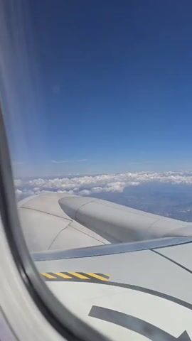 This is a video taken from the window of an airplane. The camera is looking out at the sky and the wing of the plane. The sky is a bright blue with some white clouds. The wing of the plane is white with some yellow and black markings. The video is taken in daylight.