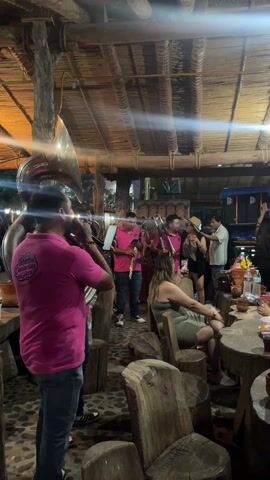The video shows a group of musicians playing brass instruments, including a tuba and trombones, in what appears to be an open-air restaurant or bar. The musicians are wearing matching pink polo shirts. The restaurant has a rustic decor with wooden furniture and a thatched roof. Several patrons are seated at tables, some of whom are watching the band. The lighting is dim, and there are some bright lights in the background. One woman sitting at a table appears to be using her cell phone. In the background, some people are standing around and talking.