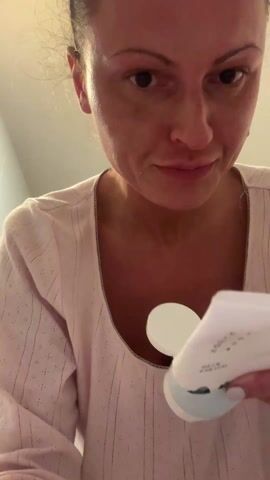 A woman in a pink and white striped shirt stands in front of a light-colored wall. She holds a white and blue tube in her right hand. The tube appears to be a skincare product, with black text written on the side. She is also holding a white applicator. Her left hand is raised with her index finger pointing upwards, as she speaks to the camera. She is looking at the camera while she talks about the product, mentioning "triple hyaluronic acid" and "ceramide NP." Her nails are painted white. The background is simple and out of focus.