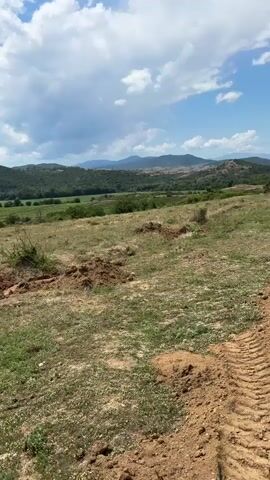 The video shows a landscape with rolling hills and mountains in the distance under a partly cloudy sky. The foreground is a dirt path with tire tracks, suggesting recent vehicle activity. There are patches of dry grass and exposed soil. In the middle ground, there are fields and scattered trees. A tall telecommunication tower is visible on a distant hilltop. The video pans from left to right, offering a wider view of the terrain.