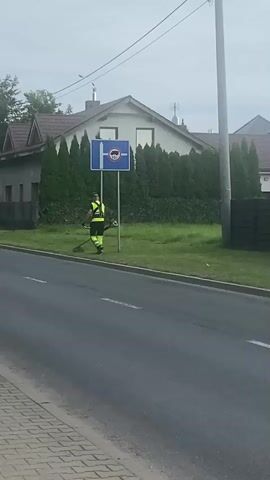 A person wearing a bright yellow and green reflective vest and green pants is trimming the grass along a roadside. They are holding a string trimmer and are positioned near a blue sign with a red circle and line through a bus icon, indicating a bus no-entry zone. In the background, there is a house with a dark roof, surrounded by tall green hedges. Another person, also in a reflective vest, can be seen further down the road, also trimming grass. The road is paved with asphalt, and a brick sidewalk is visible on the left side. The sky is overcast.