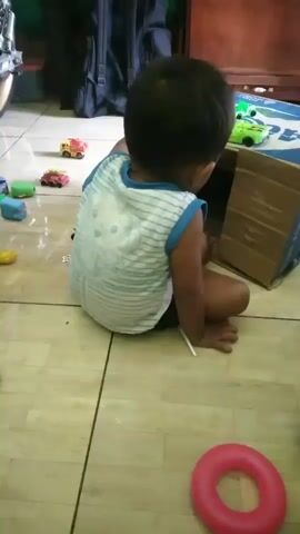 A toddler sits on a tiled floor, facing away from the camera, engaged with toys. The child is wearing a white and blue striped tank top and dark shorts. Scattered around are various toy cars, a pink ring, and a cardboard box. In one segment, the toddler turns to smile at the camera. The environment appears to be an indoor space with wooden furniture and a motorcycle visible in the background.