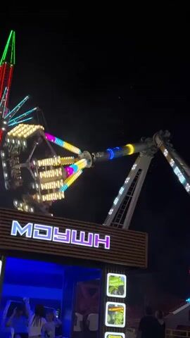 The video shows a carnival ride at night. The ride has a large, A-frame structure with lights and moving parts. People are visible in the foreground and on the ride. The ride appears to be in motion, with the arms swinging and rotating. The background is dark, indicating that it is nighttime. The overall scene suggests a lively atmosphere at a carnival or amusement park.