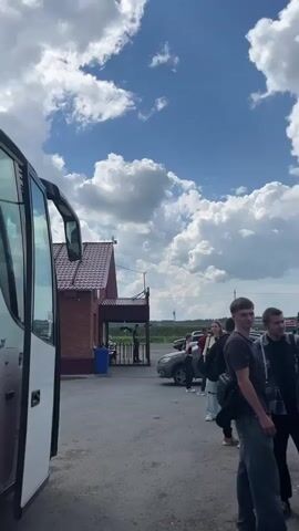A white bus is parked with its door open. Several people, including young adults and a male with short brown hair, stand on a paved area in front of a brick building. A gray car is parked behind them. The sky is blue with white clouds.