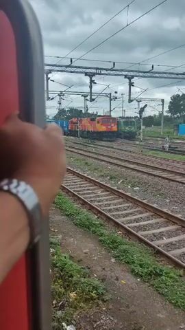 The video opens with a close-up shot of a person's hand gripping the side of a train car, with a watch on their wrist. The perspective shifts to an external view of the train, showing multiple railway tracks stretching into the distance. Several trains, including locomotives in red, yellow, and blue, are parked on adjacent tracks under an overcast sky. The environment appears to be a train yard or station, with overhead electrical lines for the trains visible. Green vegetation lines the sides of the tracks, with some litter scattered around. The camera pans slightly, showing the moving train passing by the stationary trains.