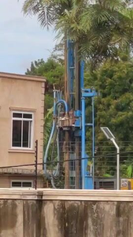 The video shows a blue drilling rig standing tall behind a concrete wall. Beside the rig is a beige building with a white-framed window and a barbed wire fence in front. A solar street light is also visible near the rig, and lush green trees and a palm tree surround the area. The sky above is overcast, and distant vehicular noise can be heard in the background.