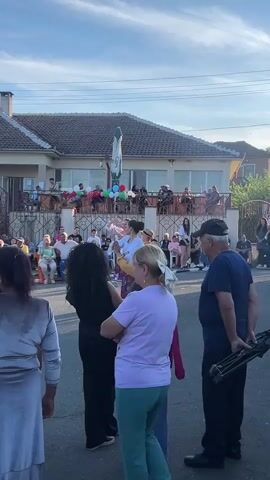 A group of people are gathered on a street in front of a two-story building. The building has a patio area with tables and chairs where more people are seated. Balloons in red, white, and green are attached to a railing on the patio. In the foreground, a group of people are watching a performance. Two women and one man are dancing in traditional clothing. The women are wearing white blouses with black bodices and long, patterned skirts. The man is wearing a yellow shirt with embroidery and a hat. The dancers are performing in the street, and the audience is standing and sitting on the side. The sun is setting, casting a warm glow on the scene. There is a white umbrella in front of the building. A man in the foreground is holding a tripod with what appears to be a camera or microphone attached.