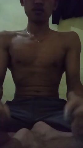 The video features a shirtless person with dark skin, sitting cross-legged with their arms bent and hands in fists. They appear to be flexing their muscles, with prominent veins visible in their forearms. The person is wearing dark shorts. The background is a plain, light-colored wall, suggesting an indoor setting. The lighting is dim, casting shadows and highlighting the person's physique. The time of day is indeterminate, but the low light suggests it could be evening or nighttime. No text is visible in the video. The overall impression is one of posing or showcasing physical strength.