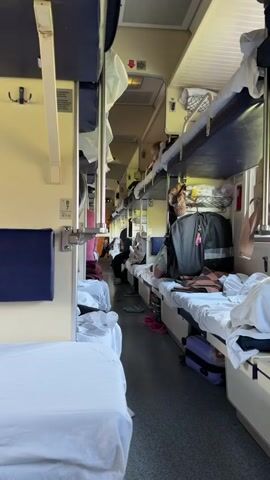 The video shows the interior of a train car. Beds are lined up on both sides of a narrow aisle. Some passengers are sitting or lying on the beds, some are on the top bunk, others on the bottom. There are personal belongings and luggage scattered around the beds. The lighting is bright. The train seems to be in motion.